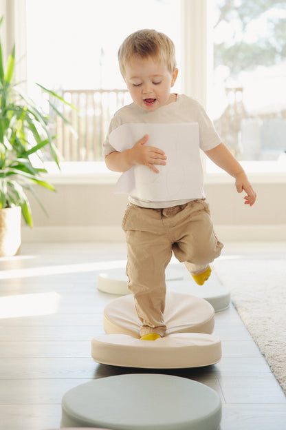 Glad dreng i gule strømper balancerer på Trollefant Balancepuder i beige og grå i lys stue, mens han holder en tegning
